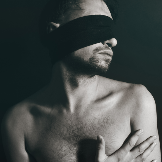 black and white photo of shirtless skinny man blind folded, arms folded across his chest, looking to his right and facing us.